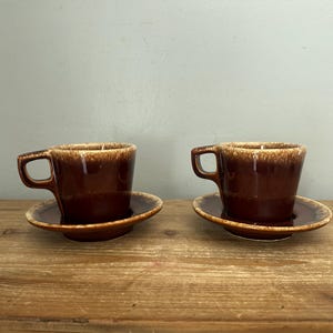 Op de afbeelding: Twee bijpassende keramische kopjes en schoteltjes met een diepbruine glazuurlaag en een lichter, gespikkeld beige druppelpatroon langs de bovenranden. Elk kopje heeft een stevig, hoekig oortje en staat in het midden op het bijbehorende schoteltje.