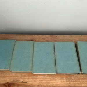 May include: Five vintage books with light blue covers are arranged on a wooden surface. The books are closed and appear to be of similar size and style, suggesting a collection or set. The covers have a textured appearance.