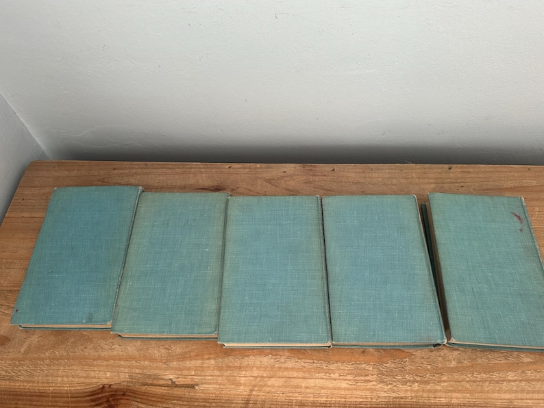 May include: Five vintage books with light blue covers are arranged on a wooden surface. The books are closed and appear to be of similar size and style, suggesting a collection or set. The covers have a textured appearance.