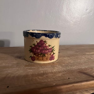 May include: A small, cream-colored ceramic pot with a blue rim and a painted design of purple flowers in a basket.