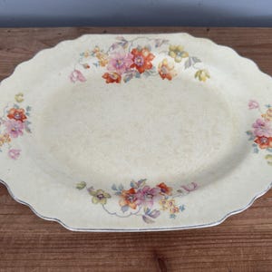 May include: Oval serving platter with a scalloped edge. The off-white platter is decorated with floral arrangements in shades of pink, orange, yellow, and green. The rim has a silver trim. A vintage piece for serving food.