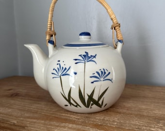 May include: White ceramic teapot with blue floral design and a woven rattan handle. The teapot has a rounded body, a spout, and a lid with a blue knob. The floral design includes blue flowers and green leaves. The handle is made of woven rattan.