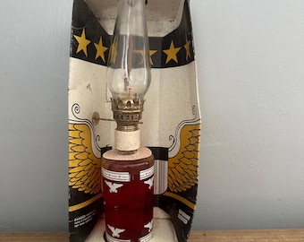 May include: Vintage oil lamp in its original box. The lamp features a clear glass chimney, a red base with white eagle designs, and a brass burner. The box is decorated with stars, eagle wings, and the word "EAGLE". The brand name "by LAMPLIGHT" is printed on the box.