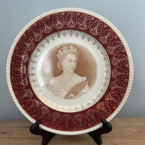 May include: A round ceramic commemorative plate featuring a sepia-toned portrait of Queen Elizabeth II in the centre. The wide border is deep red with gold leaf leaf-patterned accents and a scalloped, beaded white edge trim.