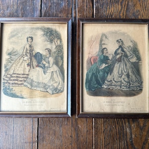 May include: Two framed vintage prints featuring women in elegant dresses. The prints, with dark wood frames, depict scenes from the past, with text at the bottom. The artwork is on aged paper, adding to the antique aesthetic.