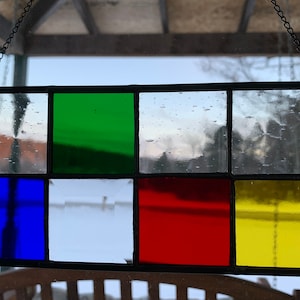 May include: A stained glass window hanging on a chain with six squares of glass. The top row has clear glass with a slight blue tint, green glass, and clear glass. The bottom row has blue glass, clear glass, red glass, and yellow glass.