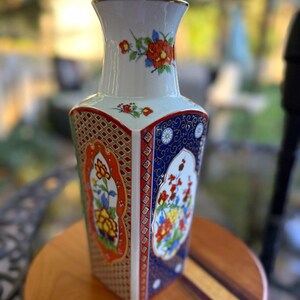 May include: A square-shaped porcelain vase featuring a white neck with floral accents and a body decorated with alternating panels of orange and blue patterns. The panels contain intricate floral motifs and geometric designs. A ruler shows a width of 7 inches.