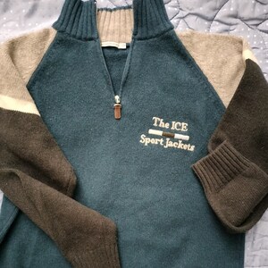 Op de afbeelding: Een teal, bruine en beige trui met een kwartrits. De mouwen zijn bruin en beige, met een witte streep op één mouw. De tekst "The ICE Sport Jackets" is op de voorkant geborduurd.