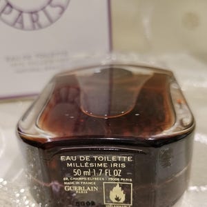 May include: A brown and clear perfume bottle with the text "EAU DE TOILETTE MILL&Eacute;SIME IRIS 50 ml 1.7 FL OZ" and the Guerlain logo. The bottle has a unique shape and a clear cap. The label also includes the text "INFLAMMABLE FLAMMABLE".