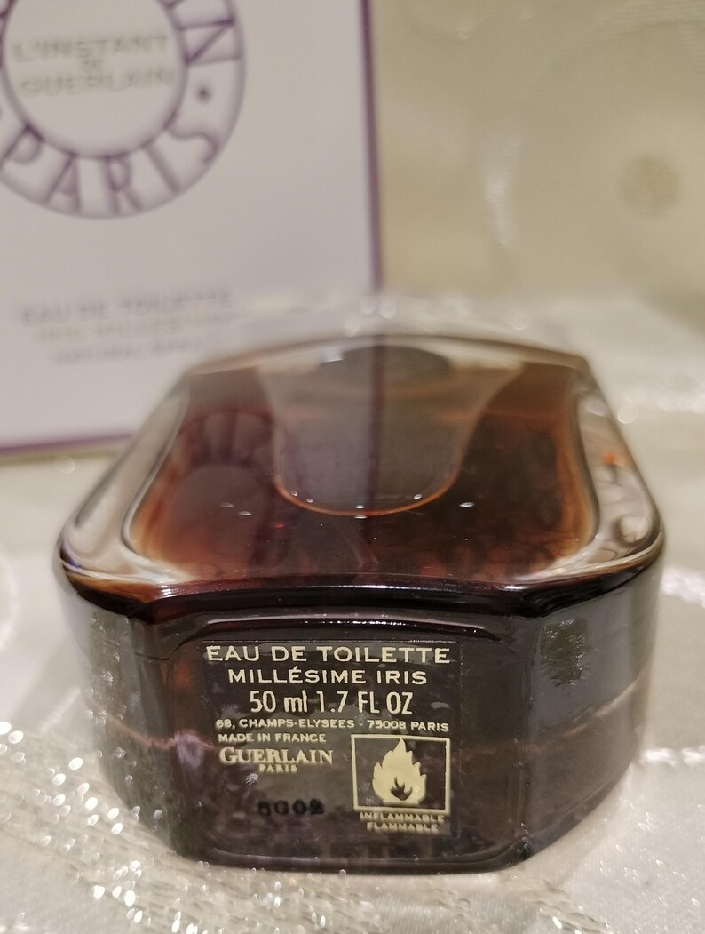 May include: A brown and clear perfume bottle with the text "EAU DE TOILETTE MILL&Eacute;SIME IRIS 50 ml 1.7 FL OZ" and the Guerlain logo. The bottle has a unique shape and a clear cap. The label also includes the text "INFLAMMABLE FLAMMABLE".