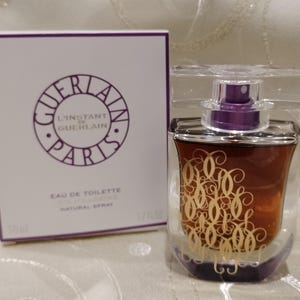 May include: A perfume bottle and box set. The bottle is a clear, curved design with a purple atomizer and a gold script design. The box is white with the words "GUERLAIN PARIS" in a purple circle.