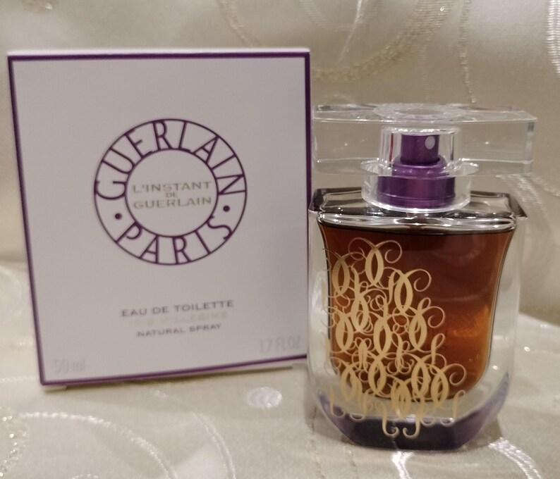 May include: A perfume bottle and box set. The bottle is a clear, curved design with a purple atomizer and a gold script design. The box is white with the words "GUERLAIN PARIS" in a purple circle.