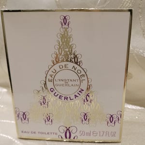May include: A white perfume box with gold accents and the text "EAU DE NOEL L'INSTANT DE GUERLAIN". The box also has decorative gold and purple designs. The bottle contains 50 ml of perfume, which is equivalent to 1.7 FL OZ.