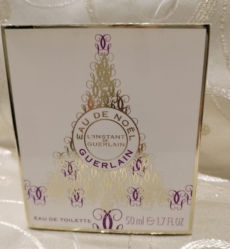 May include: A white perfume box with gold accents and the text "EAU DE NOEL L'INSTANT DE GUERLAIN". The box also has decorative gold and purple designs. The bottle contains 50 ml of perfume, which is equivalent to 1.7 FL OZ.