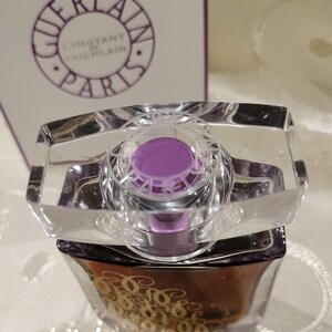 May include: A perfume bottle with a clear, rectangular top and a purple cap. The bottle has a dark brown base with gold lettering. The box in the background displays the words "GUERLAIN L'INSTANT DE GUERLAIN PARIS".