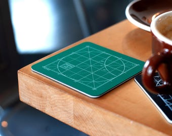 May include: A green circular protractor coaster with white lines and numbers. The coaster is on a wooden table next to a brown coffee cup and a black coaster with a geometric design.