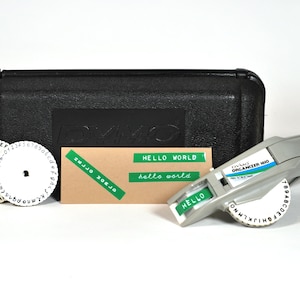 Puede incluir: Una rotuladora Dymo Organizer 1610 gris con una rueda de caracteres blanca, dos ruedas de caracteres blancas adicionales y un estuche negro. Una tarjeta beige muestra etiquetas adhesivas verdes con el texto HELLO WORLD y hello world.