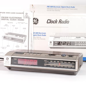 Peut inclure: Un radio-réveil numérique électronique FM/AM GE vintage avec un affichage LED rouge indiquant 3h08. Le radio-réveil a un boîtier argenté et marron, avec un cordon d'alimentation noir et une boîte en arrière-plan.