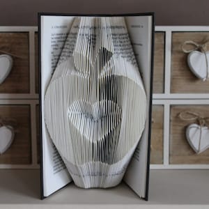 May include: A book folded into the shape of an apple. The book is standing upright on a shelf with a set of drawers behind it. The drawers are made of wood and have white trim. Each drawer has a small white heart hanging from it.