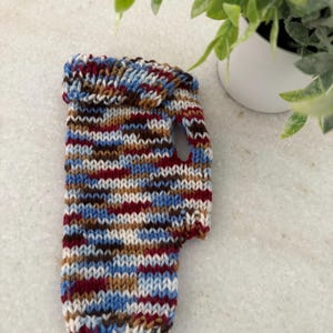 May include: A hand-knitted mug cozy in a mix of colors including blue, red, brown, and white. The cozy has a handle and is designed to fit snugly around a standard-sized mug. The cozy is made of soft yarn and provides insulation.