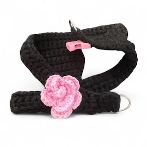 May include: Black crochet harness with a pink flower accent. The harness features two silver metal rings and a pink plastic clasp. The flower is a light pink color and is attached to the front of the harness.