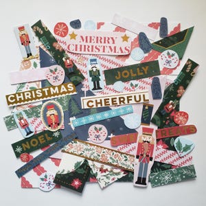 May include: A collection of Christmas-themed paper cutouts and embellishments. Includes rectangular labels with text like "Merry Christmas", "Jolly", and "Cheerful". Features nutcracker figures, snowflakes, and festive patterns in red, green, and blue.