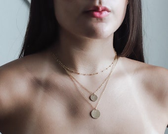 May include: A woman wearing a layered gold necklace with two circular pendants. The necklace is made of delicate chains.