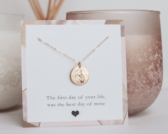 May include: Gold necklace with a round pendant featuring a line drawing of a mother holding a baby. The card reads "The first day of your life, was the best day of mine".