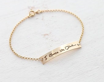Puede incluir: Un brazalete de cadena dorada con un dije de barra curvado grabado con el mensaje escrito a mano "I Choose Us, I Do".