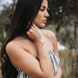 Puede incluir: Una pulsera dorada con un colgante de cruz y una peque&ntilde;a gema roja. La pulsera se lleva en la mu&ntilde;eca. La persona lleva un top sin tirantes a rayas blancas y negras.