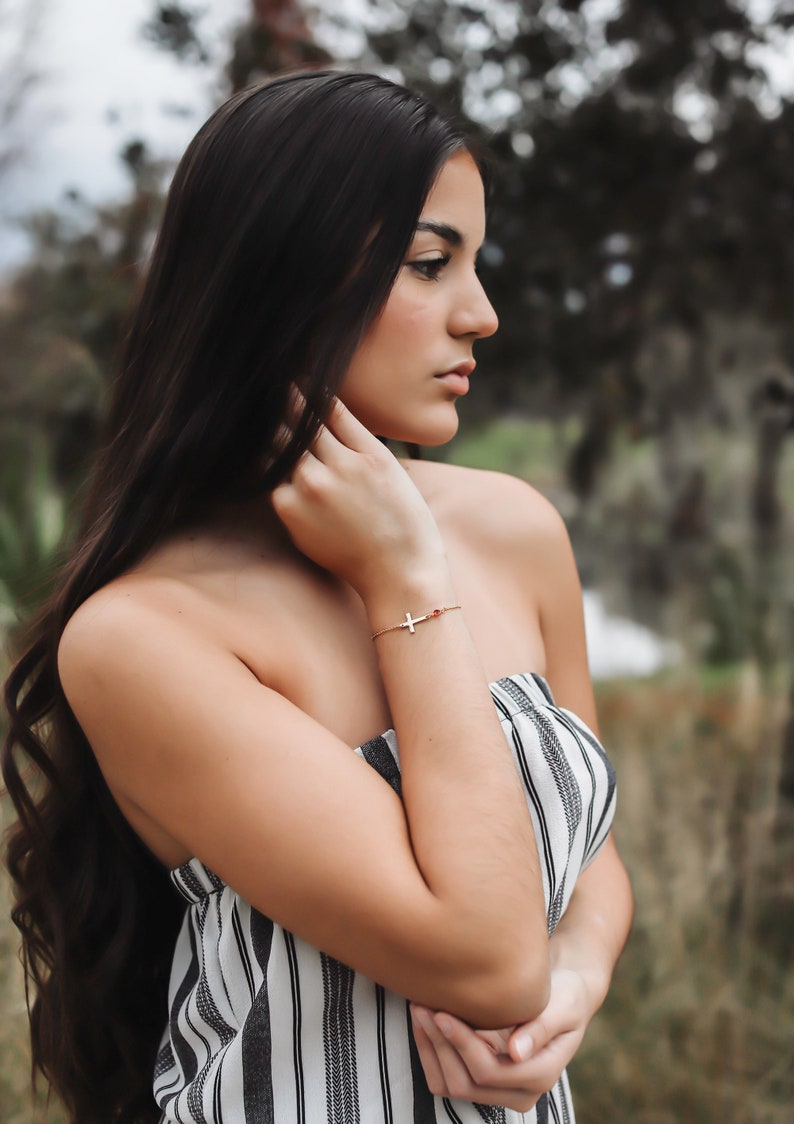 Puede incluir: Una pulsera dorada con un colgante de cruz y una peque&ntilde;a gema roja. La pulsera se lleva en la mu&ntilde;eca. La persona lleva un top sin tirantes a rayas blancas y negras.
