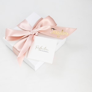 May include: A white gift box with a pink satin ribbon tied in a bow. The box has a white label with the text "Reflection of memories" and a pink ribbon with the name "Taylor" written in gold lettering.