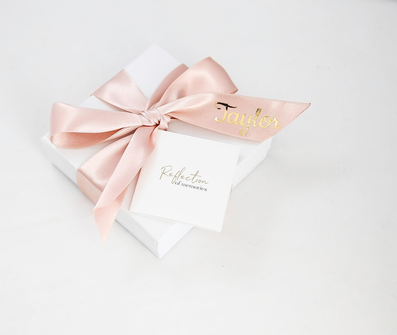 May include: A white gift box with a pink satin ribbon tied in a bow. The box has a white label with the text "Reflection of memories" and a pink ribbon with the name "Taylor" written in gold lettering.