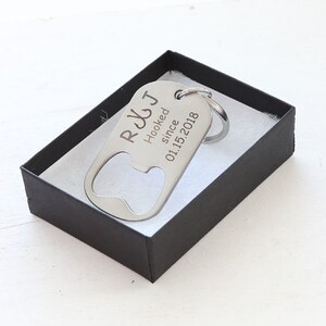 May include: A silver bottle opener keychain with the text "R&J Hooked since 01.15.2018" is displayed inside a black gift box with white lining. The keychain has a bottle opener on one end and a key ring on the other.