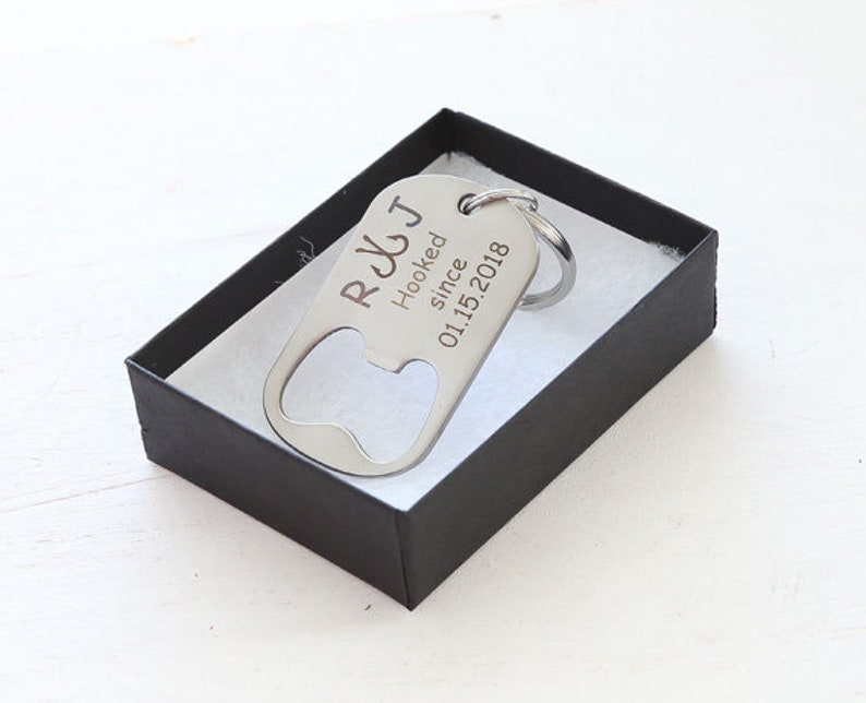 May include: A silver bottle opener keychain with the text "R&J Hooked since 01.15.2018" is displayed inside a black gift box with white lining. The keychain has a bottle opener on one end and a key ring on the other.