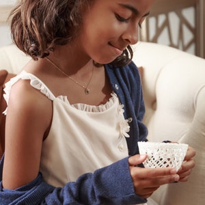 May include: A young girl wearing a white dress with a blue cardigan and a gold necklace with a green stone pendant. She is holding a white crocheted cup.