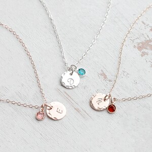May include: Three delicate gold necklaces with small round pendants featuring a single letter and a tiny gemstone. The necklaces are on a white wooden surface.