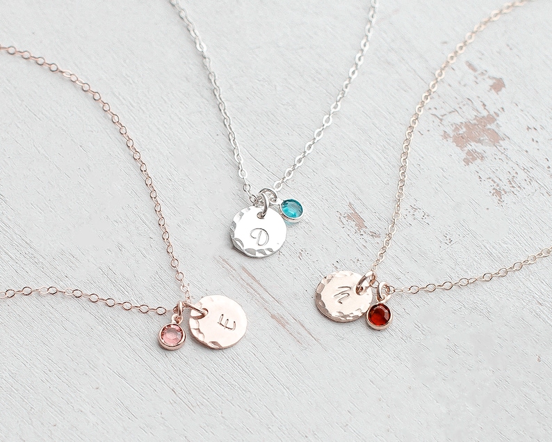 May include: Three delicate gold necklaces with small round pendants featuring a single letter and a tiny gemstone. The necklaces are on a white wooden surface.