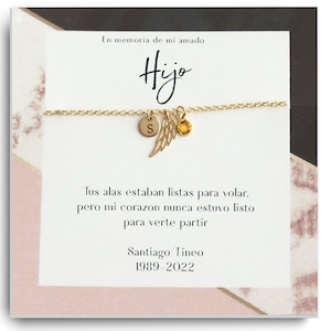 Op de afbeelding: Een gouden kettingarmband met een kleine engelenvleugelbedel en een letter 'S'-bedel. De armband wordt getoond op een kaart met de tekst "En memoria de mi amado Hijo" en een gedicht in het Spaans. De kaart bevat ook de naam "Santiago Tineo" en de data "1989-2022".