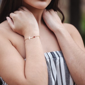 Puede incluir: Una pulsera dorada con un dije de cruz y una peque&ntilde;a cuenta roja. La pulsera se lleva en la mu&ntilde;eca. El fondo est&aacute; borroso, lo que sugiere un entorno al aire libre. La pulsera es una joya.