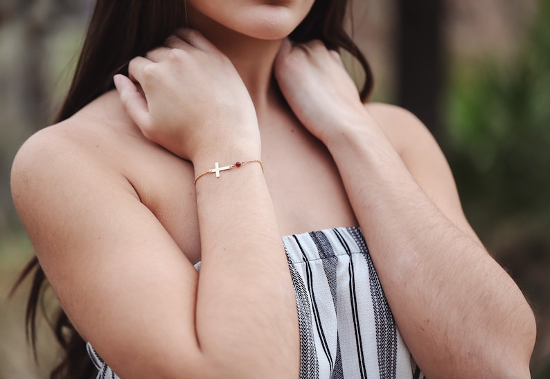 Puede incluir: Una pulsera dorada con un dije de cruz y una peque&ntilde;a cuenta roja. La pulsera se lleva en la mu&ntilde;eca. El fondo est&aacute; borroso, lo que sugiere un entorno al aire libre. La pulsera es una joya.