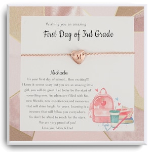 Op de afbeelding: Een roségouden armband met een hartvormige bedel met de letter "M" wordt gepresenteerd op een kaart. De kaart leest "Wishing you an amazing First Day of 3rd Grade" en bevat een bericht en een schoolthema-illustratie.