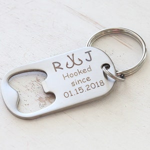 May include: A silver-colored bottle opener keychain with a rounded end and a key ring. The opener has the engraved text "R J Hooked since 01.15.2018" and two fish hook symbols.