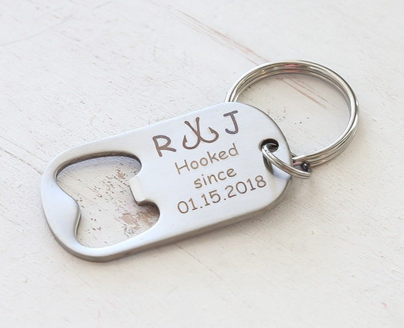 May include: A silver-colored bottle opener keychain with a rounded end and a key ring. The opener has the engraved text "R J Hooked since 01.15.2018" and two fish hook symbols.