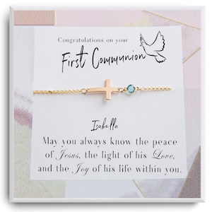 May include: A white card with a gold cross necklace and a blue gemstone. The card has the text "Congratulations on your First Communion" and "Isabella" with a dove illustration. The card also has the text "May you always know the peace of Jesus, the light of his Love, and the Joy of his life within you."