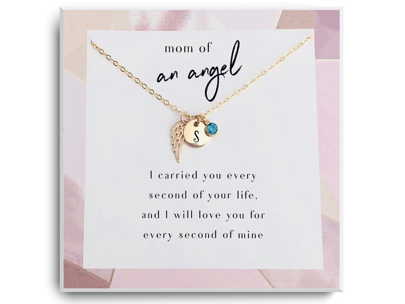 May include: A gold necklace with a small angel wing charm and a round charm with the letter "S" and a turquoise gemstone. The necklace comes in a white box with the text "mom of an angel" and "I carried you every second of your life, and I will love you for every second of mine."
