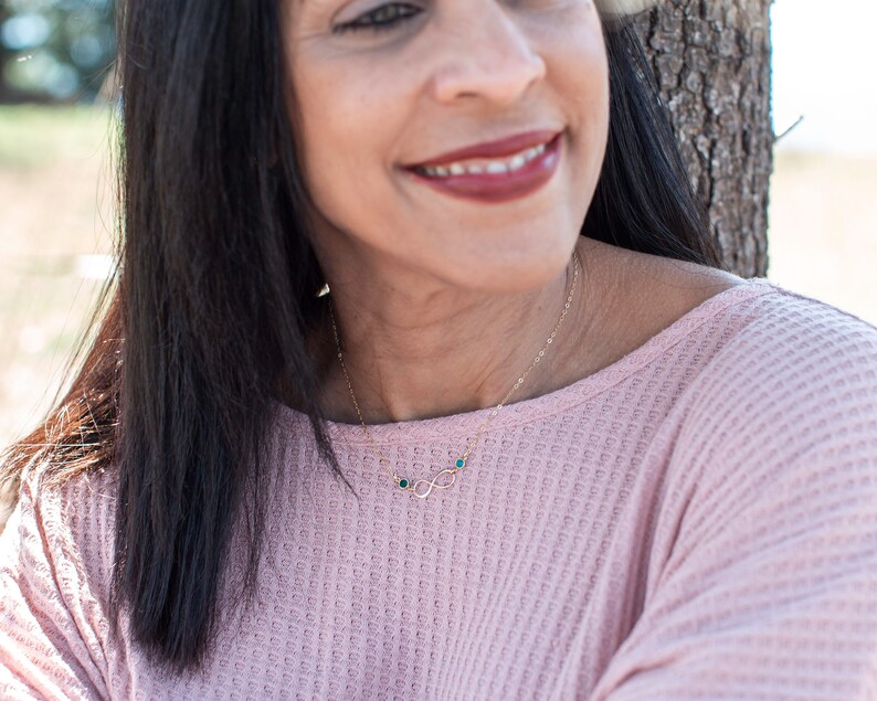 May include: A woman wearing a pink waffle knit top with a gold infinity necklace with small blue gemstones.