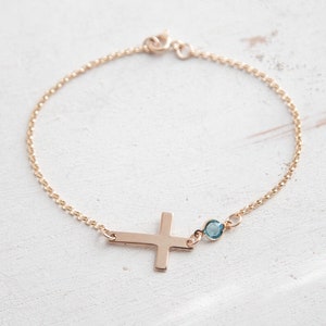 Puede incluir: Una delicada pulsera dorada con un colgante de cruz lateral y una peque&ntilde;a gema azul. La pulsera tiene una fina cadena y un cierre sencillo. La cruz es de dise&ntilde;o s&oacute;lido y plano.