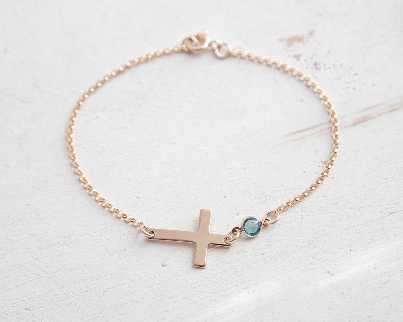 Puede incluir: Una delicada pulsera dorada con un colgante de cruz lateral y una peque&ntilde;a gema azul. La pulsera tiene una fina cadena y un cierre sencillo. La cruz es de dise&ntilde;o s&oacute;lido y plano.