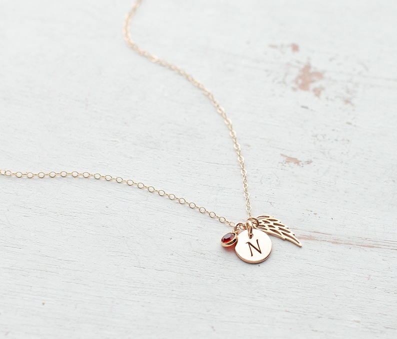 May include: Gold necklace with a small red gemstone, a charm with the letter 'N' and a delicate angel wing charm.